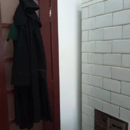 Toga adwokacka z zielonym żabotem wisząca na haczyku na ciemnoczerwonych drzwiach, obok pieca kaflowego z ozdobną kratką wentylacyjną.