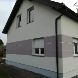 Ocieplony dom jednorodzinny z jasnoszarą elewacją i poziomym pasem w kolorze taupe, widok z boku, z widocznymi oknami z roletami zewnętrznymi.