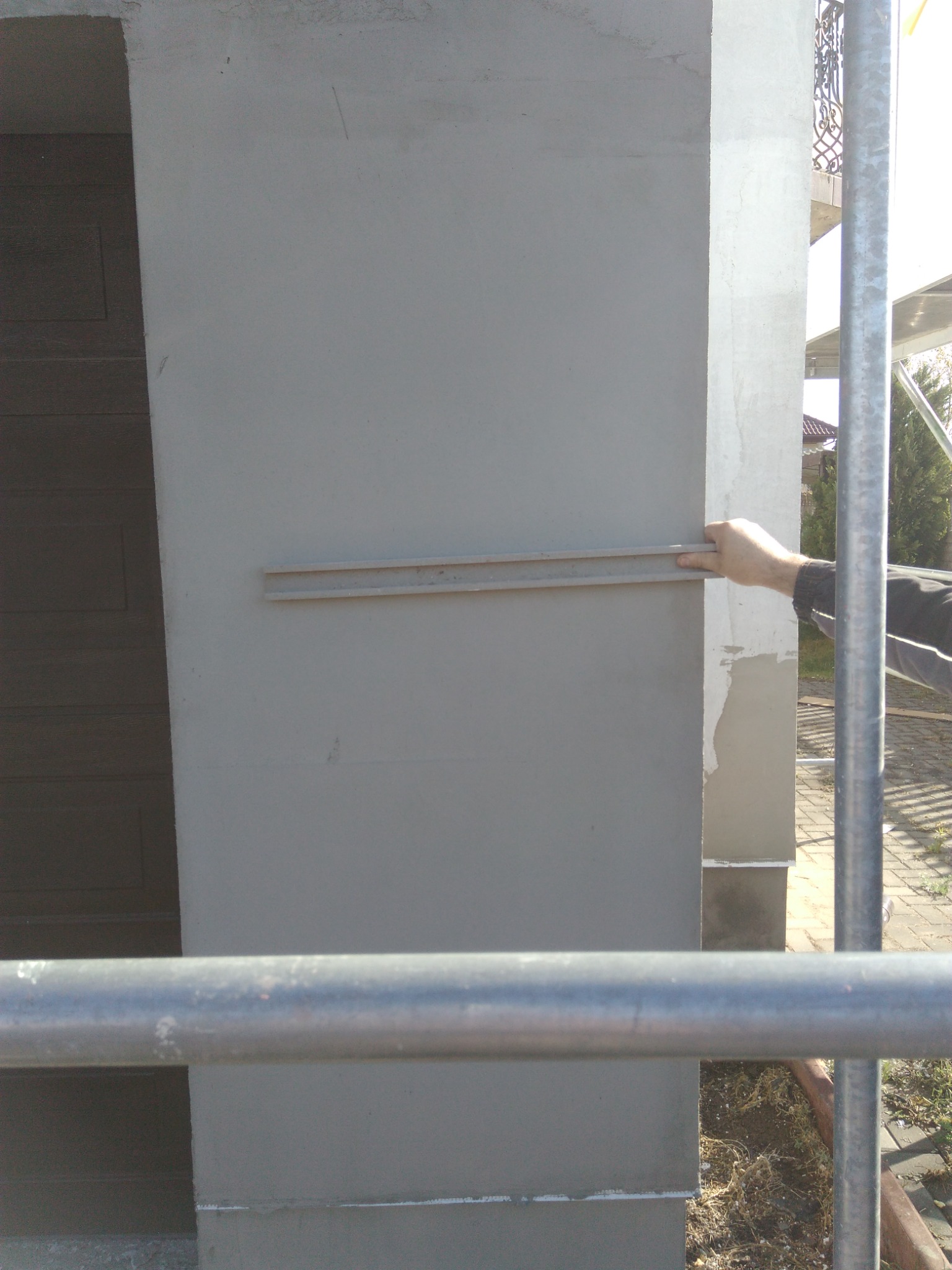 Mężczyzna wyrównuje listwą świeżo nałożony tynk na filarze elewacyjnym, widoczne rusztowanie i fragment elewacji z dekoracyjnym balkonem.