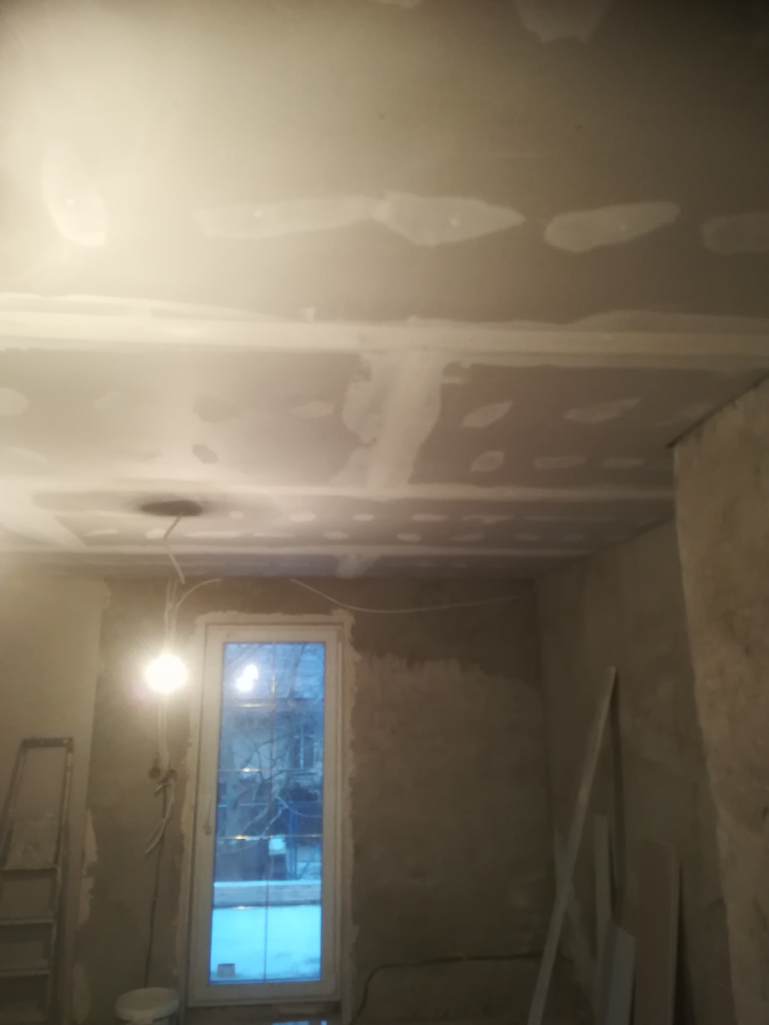 Poddasze w trakcie prac wykończeniowych, widoczna surowa powierzchnia sufitu z płyt gipsowo-kartonowych, szpachlowana, z widocznymi łączeniami, okno z widokiem na zewnątrz, drabina przystawiona...