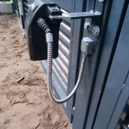 Szczegółowe ujęcie siłownika automatyki do bramy wjazdowej z widocznymi śrubami mocującymi i przewodami w metalowej osłonie, zamontowanego na metalowej, poziomej bramie lamelowej w kolorze...