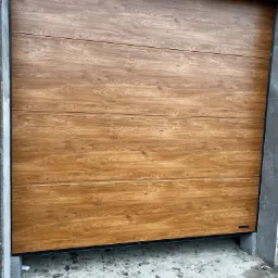 Zamknięta segmentowa brama garażowa z panelami o fakturze jasnego drewna, widok z bliska na fragment ściany garażu z surowego betonu.