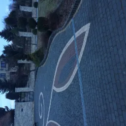 Ułożona kostka brukowa w kolorze grafitowym z dekoracyjnym wzorem liścia w odcieniach brązu i beżu, widok na dom i ogrodzenie z kamienia.