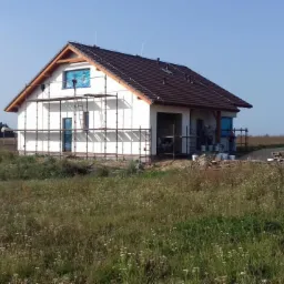 Dom jednorodzinny w trakcie prac wykończeniowych elewacji, widoczne rusztowania, biała fasada, brązowy dach, teren niezagospodarowany, w tle zieleń.