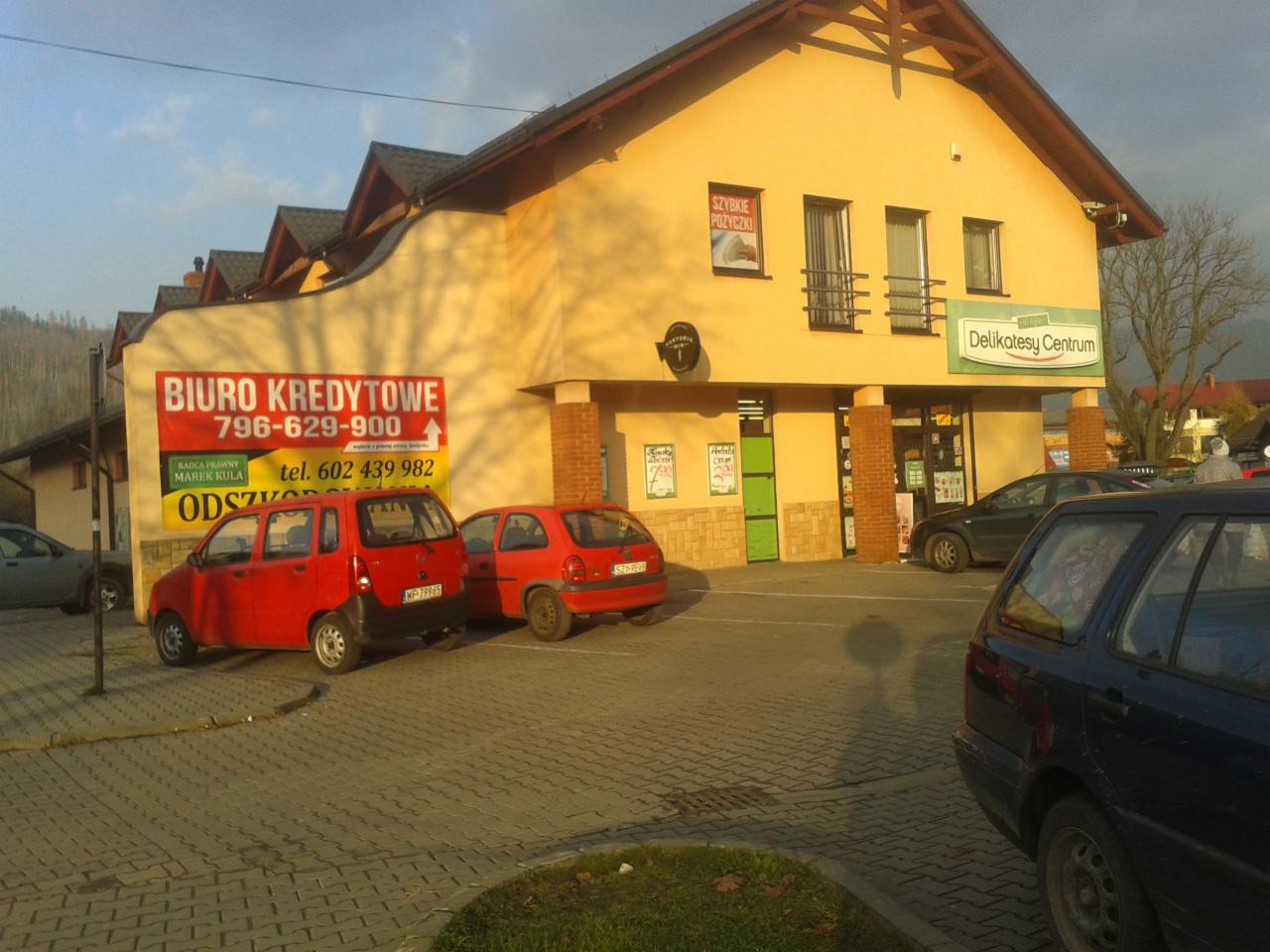 Budynek z szyldem 'Biuro Kredytowe' i 'Delikatesy Centrum', zaparkowane samochody na kostce brukowej, widok na góry w tle.