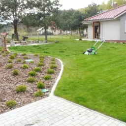 Zadbany ogród z równo przystrzyżonym trawnikiem, rabatą wysypaną korą i niskimi krzewami, oraz zieloną kosiarką na trawie przed domem z czerwoną dachówką.