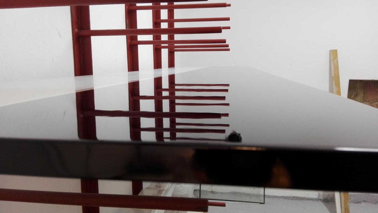 Błyszczący, czarny blat w trakcie lakierowania, wiszący na metalowym stojaku w lakierni, widoczne odbicie konstrukcji stojaka w powierzchni blatu.