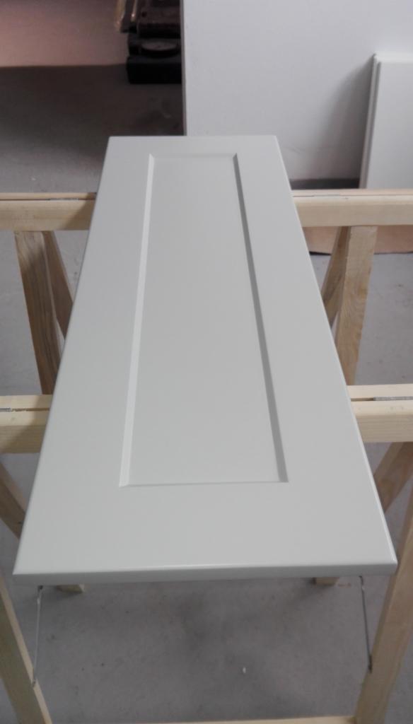 Pomalowane na szaro skrzydło drzwiowe z frezowanym wzorem, leżące na drewnianym stojaku w warsztacie.