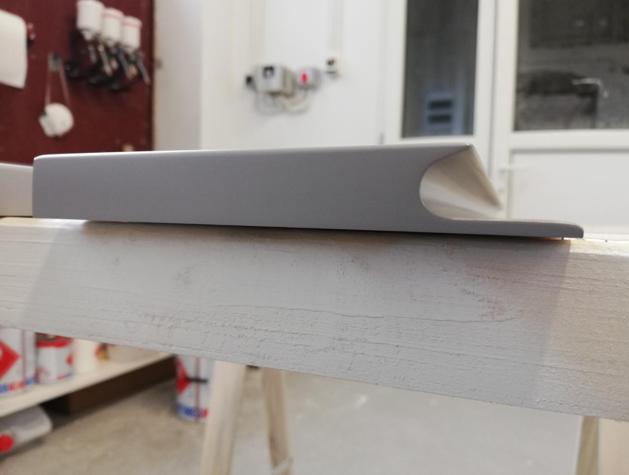 Szary, lakierowany element drewniany z zaokrąglonym brzegiem, leżący na białym, drewnianym blacie w warsztacie stolarskim, widoczne puszki z farbą i narzędzia w tle.