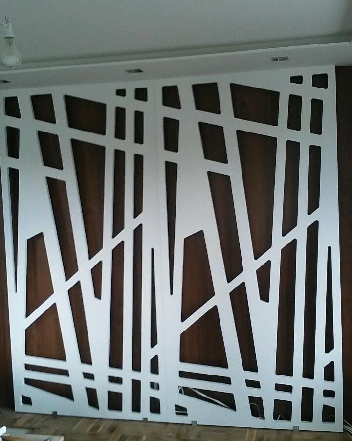 Białe, ażurowe panele dekoracyjne z geometrycznym wzorem na brązowym tle, widoczne przewody elektryczne u dołu.