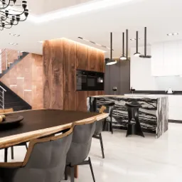 Nowoczesna kuchnia z wyspą z czarnego marmuru, drewnianą zabudową, schodami z metalowymi barierkami i dużym stołem z orzechami w misie.