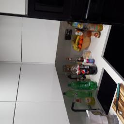 Remix - Nowoczesna kuchnia z białymi szafkami, czarnymi akcentami, blatem z widocznymi produktami spożywczymi i akcesoriami kuchennymi, takimi jak deska do krojenia i butelki z płynami.