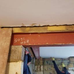 Remix - Montaż stalowej belki nośnej w trakcie remontu, widoczne fragmenty muru, sufitu i pracownicy w rękawicach ochronnych, jeden z nich trzyma białą płytę.