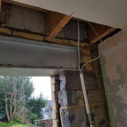 Remix - Wnętrze w trakcie prac budowlanych: stalowa belka nad otworem okiennym, fragment wymurowanej ściany z pustaków, wystające przewody elektryczne, ślady zaprawy i laserowy poziom na ścianie.