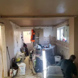 Wnętrze w trakcie prac wykończeniowych: świeżo otynkowane ściany, widoczne narzędzia, drabina, okno z widokiem na balkon, pracownik w trakcie wygładzania ściany. Część pomieszczenia zabezpieczona...