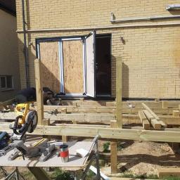 Remix - Budowa drewnianego tarasu przy domu z cegły, widoczne deski szkieletu, narzędzia i pracujący mężczyzna; drzwi balkonowe zabezpieczone płytą OSB.