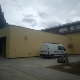 Budynek o jednolitej, jasnej elewacji z widocznym systemem monitoringu i zaparkowanym białym vanem na kostce brukowej.