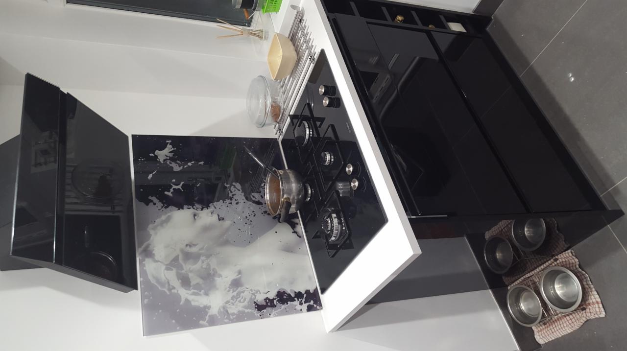 Nowoczesna kuchnia z czarnymi, lakierowanymi frontami szafek, blatem w kolorze białym, czarną płytą grzewczą i szklanym panelem nad blatem z motywem chmur.