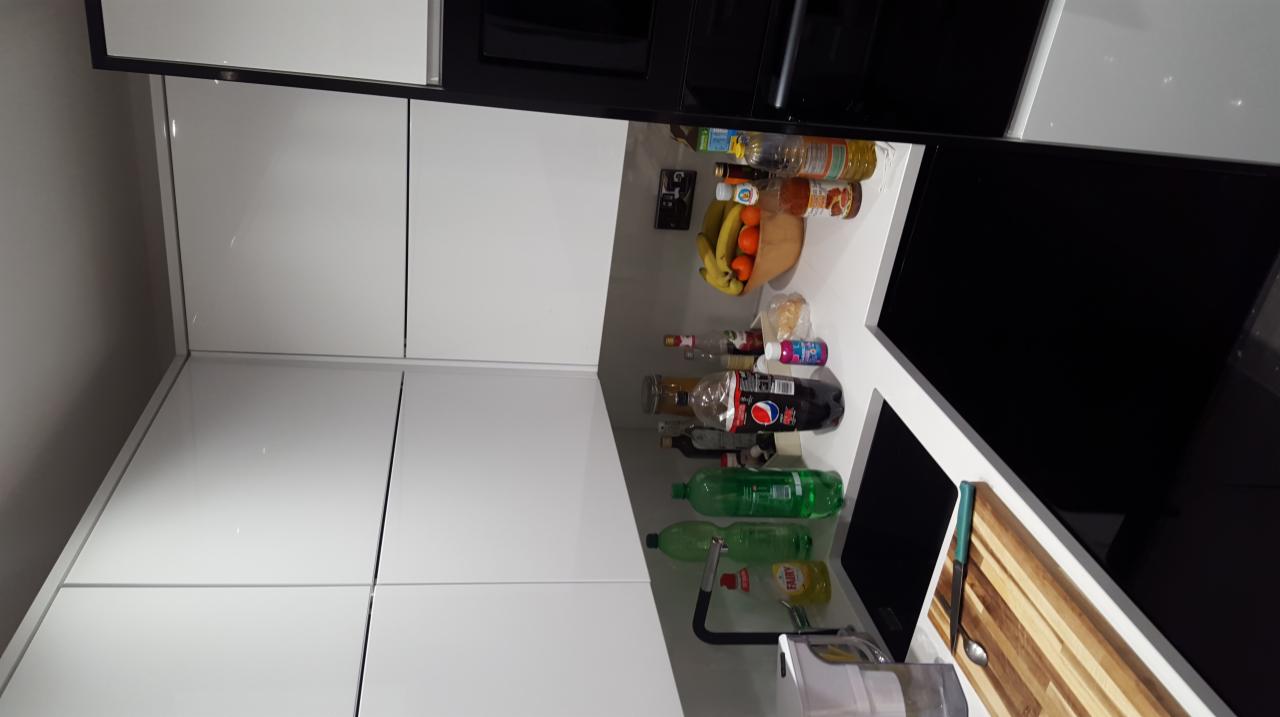 Nowoczesna kuchnia z białymi szafkami, czarnymi akcentami, blatem z widocznymi produktami spożywczymi i akcesoriami kuchennymi, takimi jak deska do krojenia i butelki z płynami.