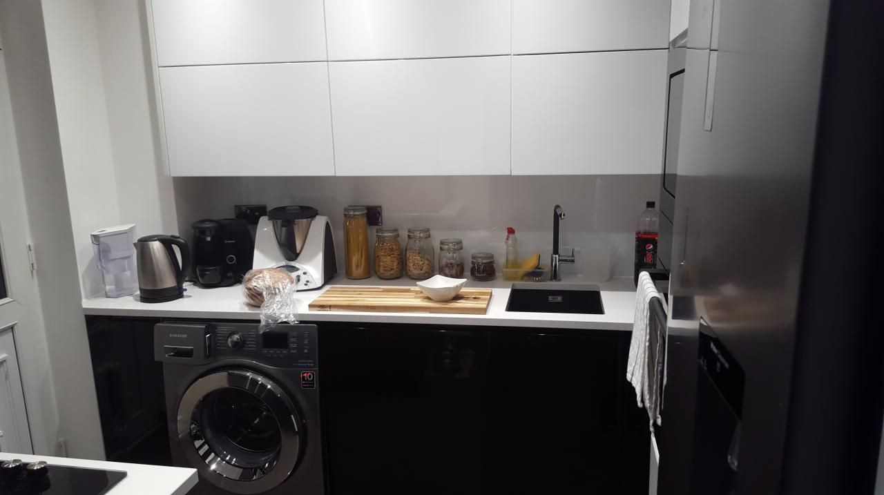 Nowoczesna kuchnia z białymi szafkami wiszącymi i czarnymi szafkami dolnymi, pralka w zabudowie pod blatem, akcesoria kuchenne na blacie, minimalistyczny styl.