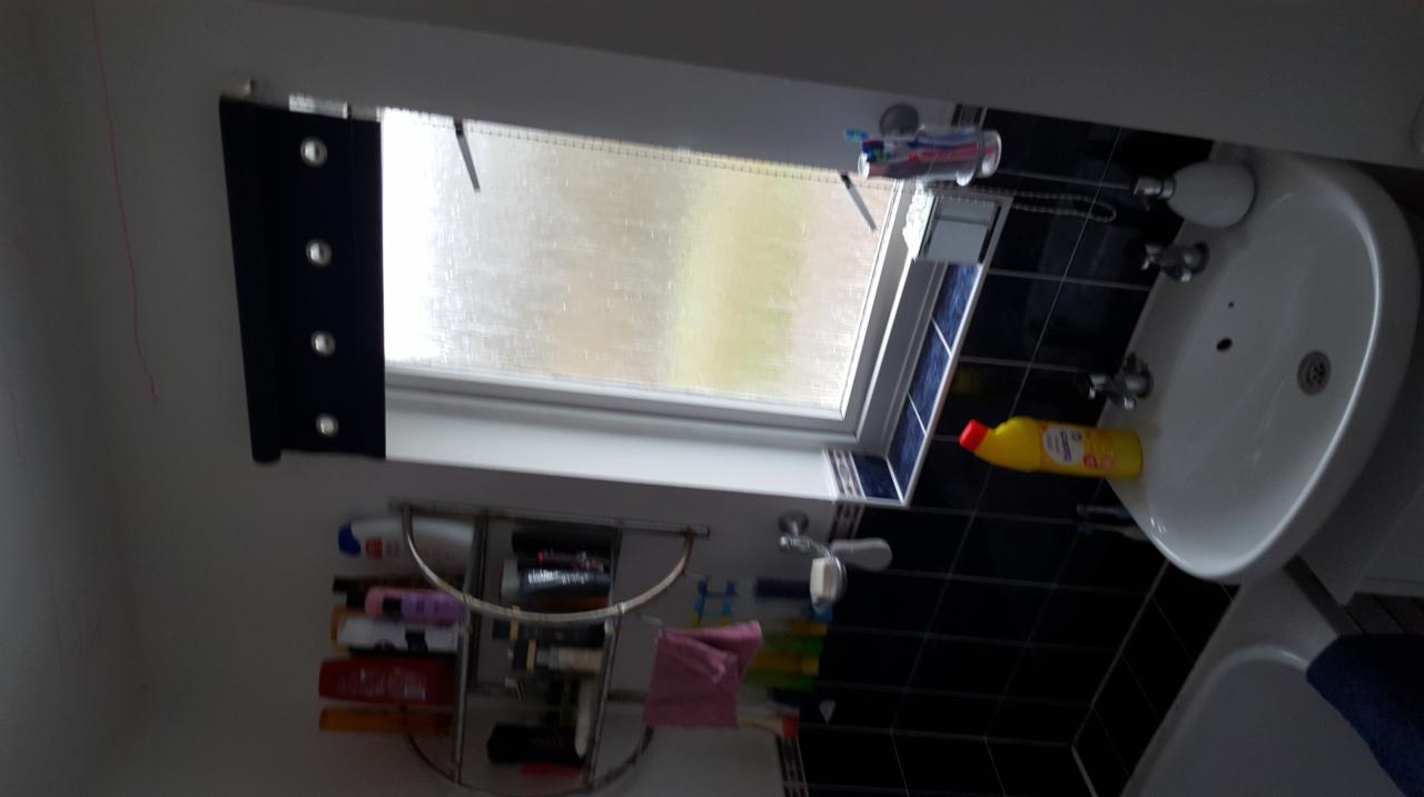 Umywalka w łazience z czarnymi płytkami, nad nią okno dachowe z roletą, widoczna butelka płynu do czyszczenia i półka z kosmetykami.