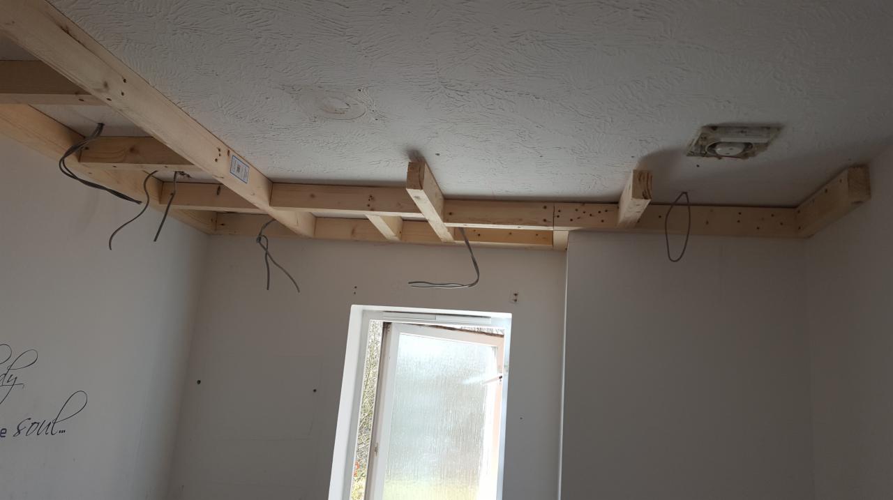 Konstrukcja drewniana podwieszanego sufitu zamontowana do sufitu z fakturą, widoczne wystające przewody elektryczne i częściowo otwarte okno.