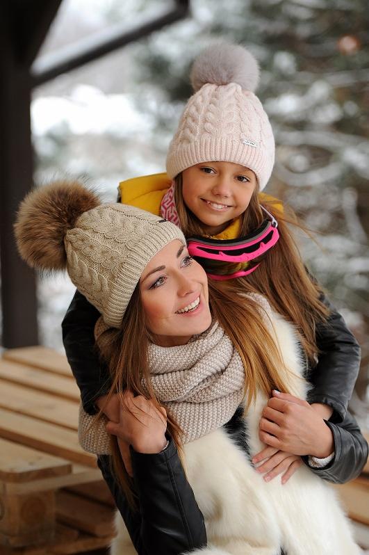 Uśmiechnięta matka i córka w pasujących dzianinowych czapkach z pomponami, na tle zimowego krajobrazu, kobieta w futrzanej kamizelce i szaliku.