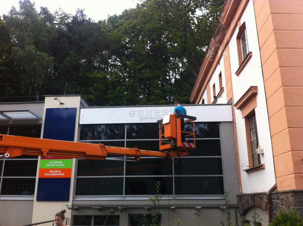 Pracownik na podnośniku montuje napis 'GENESIS STUDIO' na elewacji budynku z dużymi oknami.