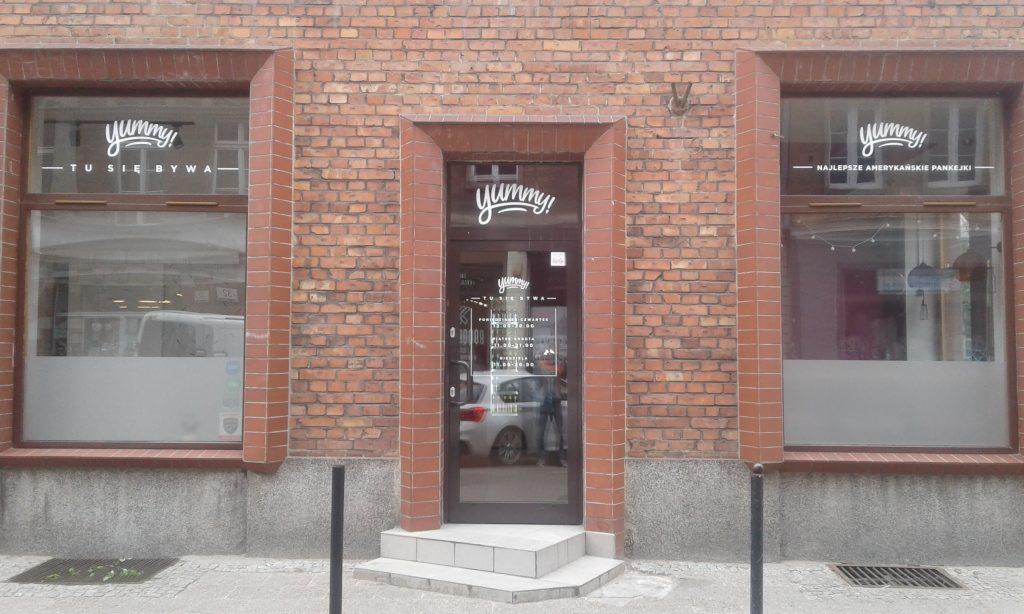 Oklejone witryny lokalu gastronomicznego z ceglaną elewacją, logo 'yummy!' i napisy informacyjne na szybach okiennych i drzwiach.