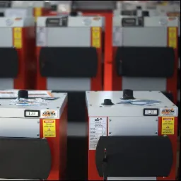 Rząd nowych, czerwono-białych pieców CO marki Kotlospaw, z widocznymi naklejkami ostrzegawczymi i danymi technicznymi, ustawionych w hali produkcyjnej lub magazynie.