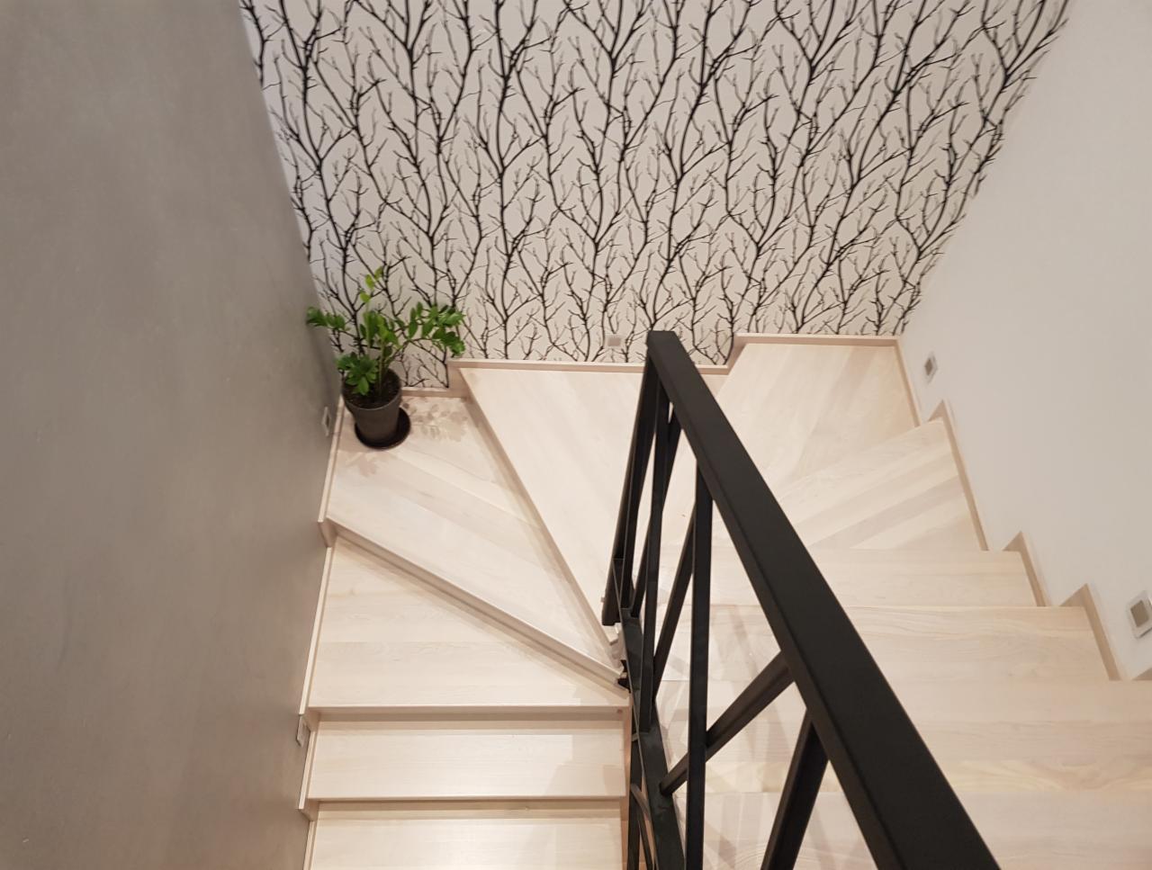Jasne drewniane schody z czarną metalową balustradą, widok z góry, ściana z tapetą w motywie gałęzi, doniczka z rośliną na pierwszym stopniu.