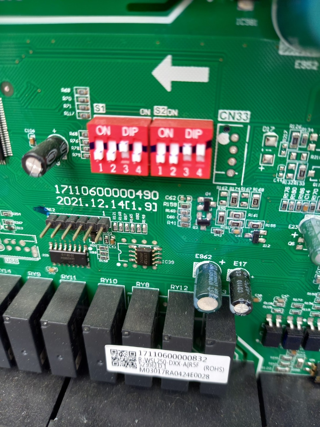 Zbliżenie na fragment zielonej płytki drukowanej z elementami elektronicznymi, w tym czerwone przełączniki DIP, kondensatory i układy scalone, z wyraźnymi oznaczeniami i numerami seryjnymi.