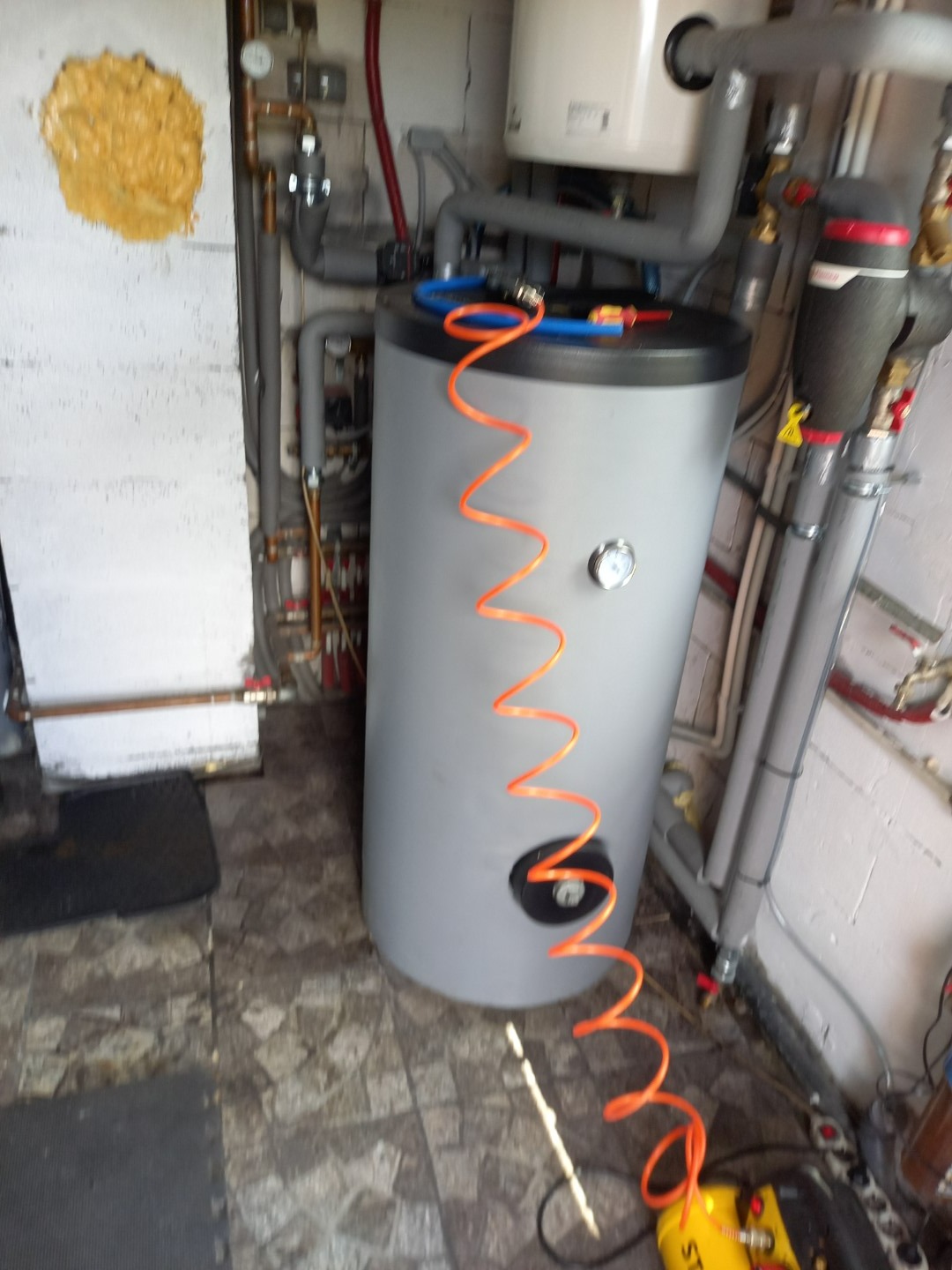 Szary zasobnik ciepłej wody użytkowej z manometrem, podłączony do instalacji grzewczej, widoczny na tle ściany z instalacjami w Warszawie. Pomarańczowy przewód sprężonego powietrza leży obok.