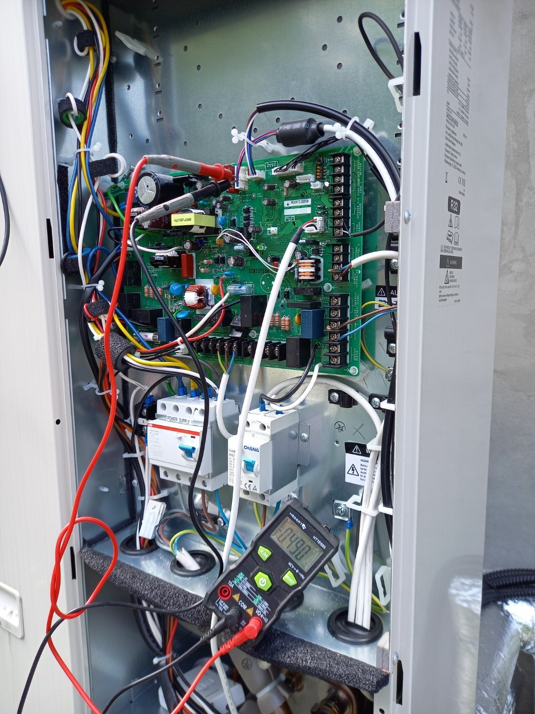 Multimetr podłączony do płyty sterującej wewnątrz obudowy urządzenia grzewczego, widoczne okablowanie, bezpieczniki i moduły elektroniczne
