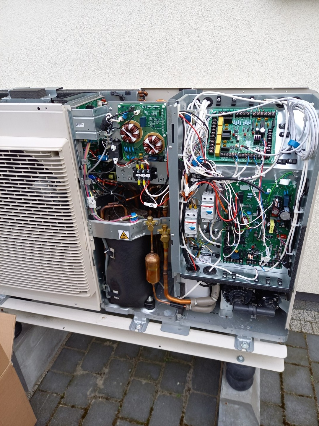Otwarta obudowa pompy ciepła z widocznymi elementami wewnętrznymi: płytki drukowane, okablowanie, transformatory i miedziane rurki, umieszczona na zewnątrz budynku.