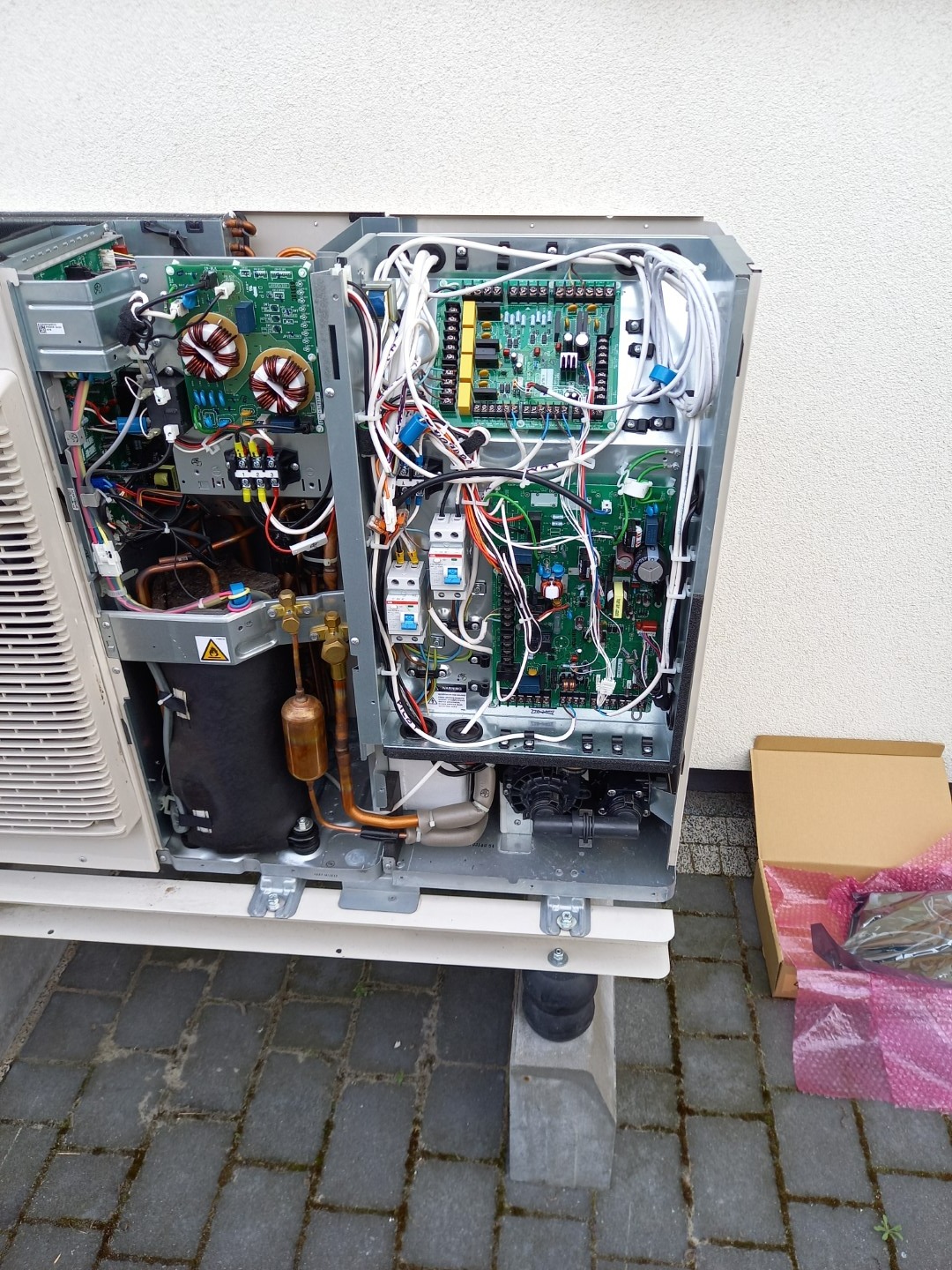 Otwarta obudowa pompy ciepła z widocznymi elementami elektronicznymi, przewodami i miedzianymi rurami, obok karton z folią bąbelkową, na zewnątrz budynku.