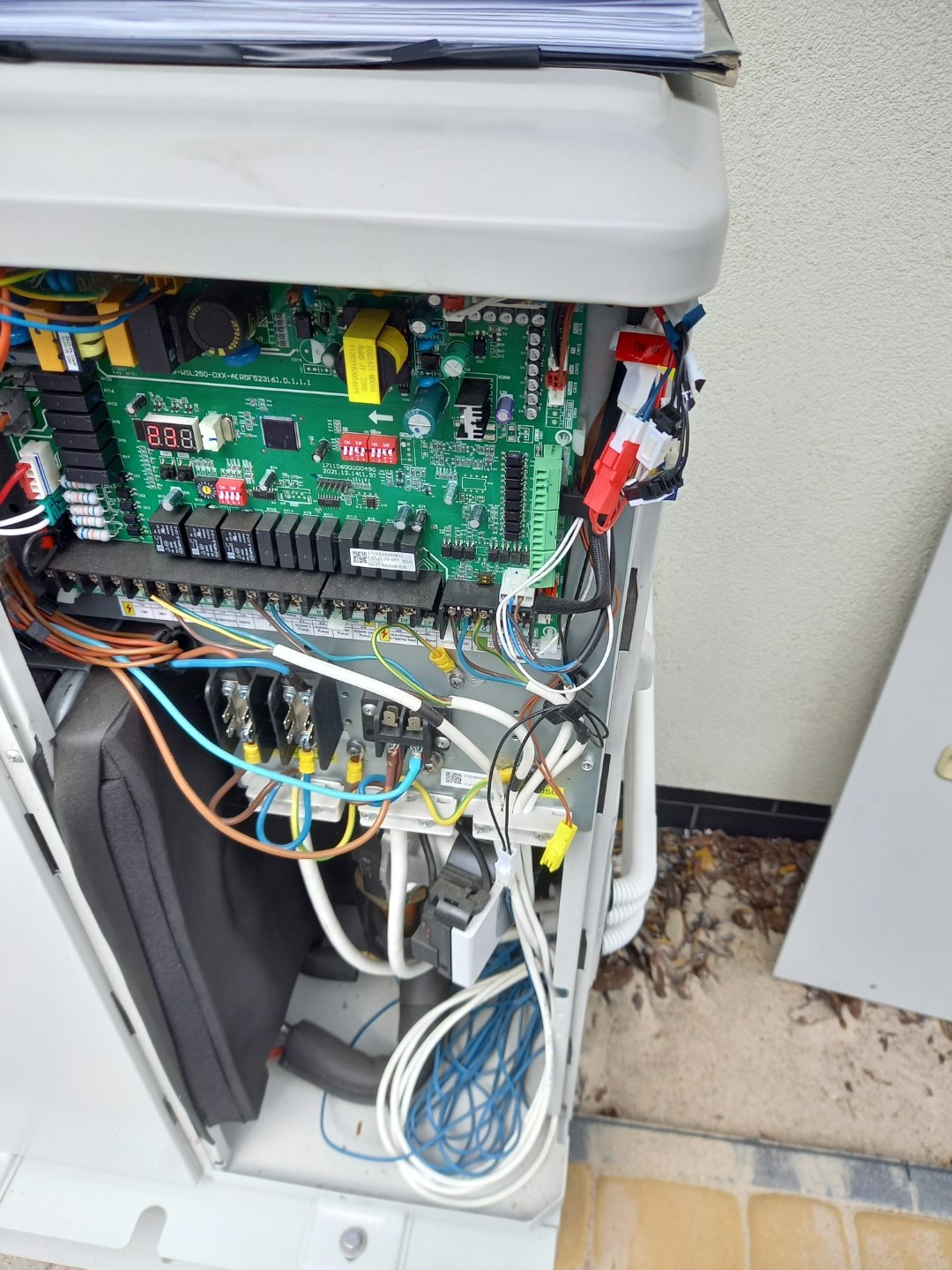 Wnętrze otwartej obudowy pieca gazowego z widoczną płytą sterującą, okablowaniem i elementami instalacji, z dokumentacją techniczną na górze.