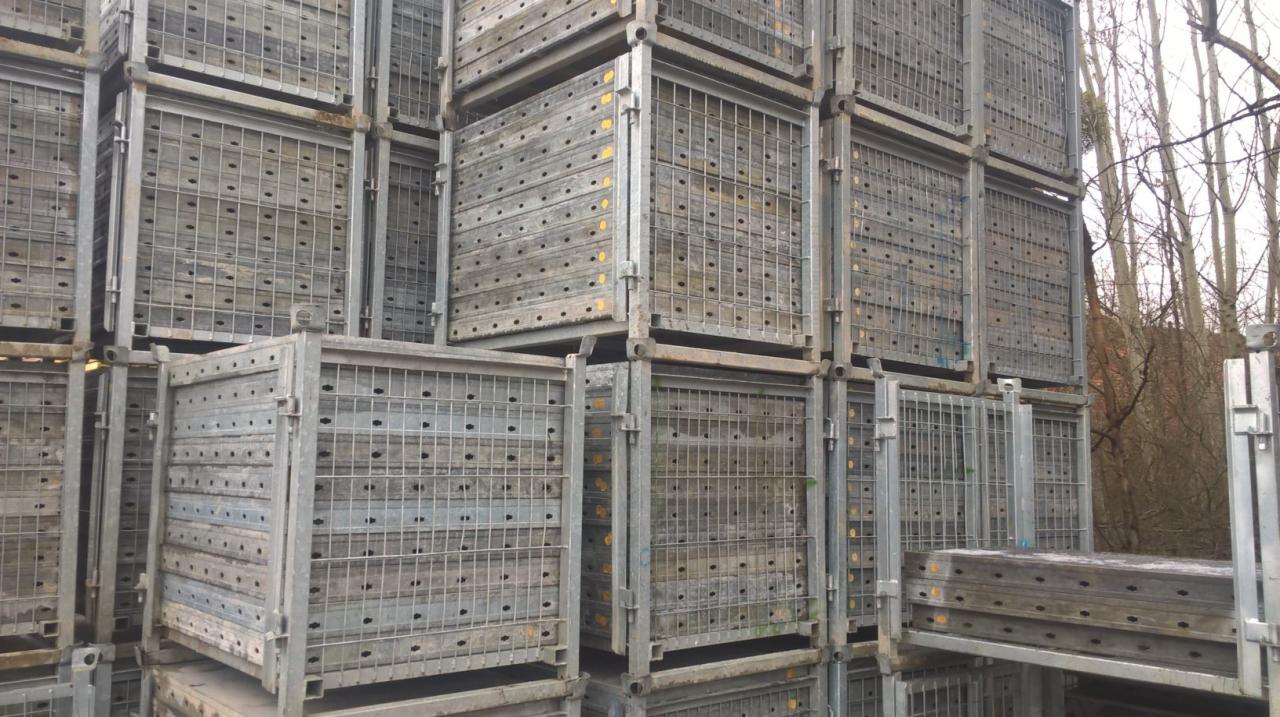 Stos metalowych koszy paletowych z drewnianymi wkładami na zewnątrz magazynu, widoczne drzewa w tle.