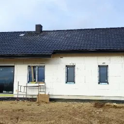 Dom jednorodzinny w trakcie budowy z zamontowanym czarnym dachem z dachówki ceramicznej i rynnami, ściany z białego bloczka, okna wstawione, teren wokół w trakcie prac ziemnych.