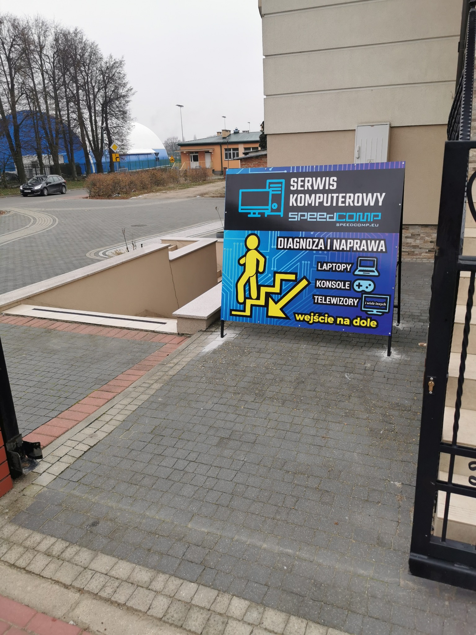 Tablica reklamowa serwisu komputerowego SpeedCOMP z ikonami laptopa, konsoli i telewizora, umieszczona przy schodach prowadzących do wejścia 'wejście na dole'.