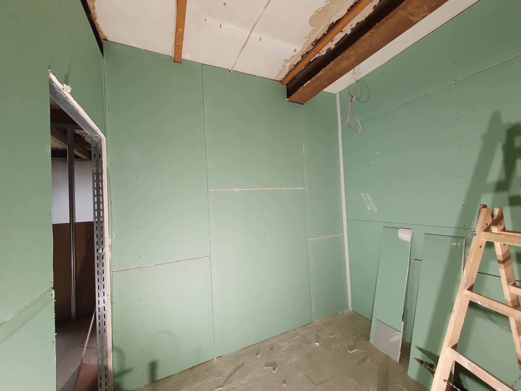 Montaż zielonych płyt karton-gips na ścianach i suficie, widoczne profile metalowe przy otworze drzwiowym, drewniana drabina oparta o ścianę, niedokończone wykończenie sufitu z odsłoniętymi belkami.