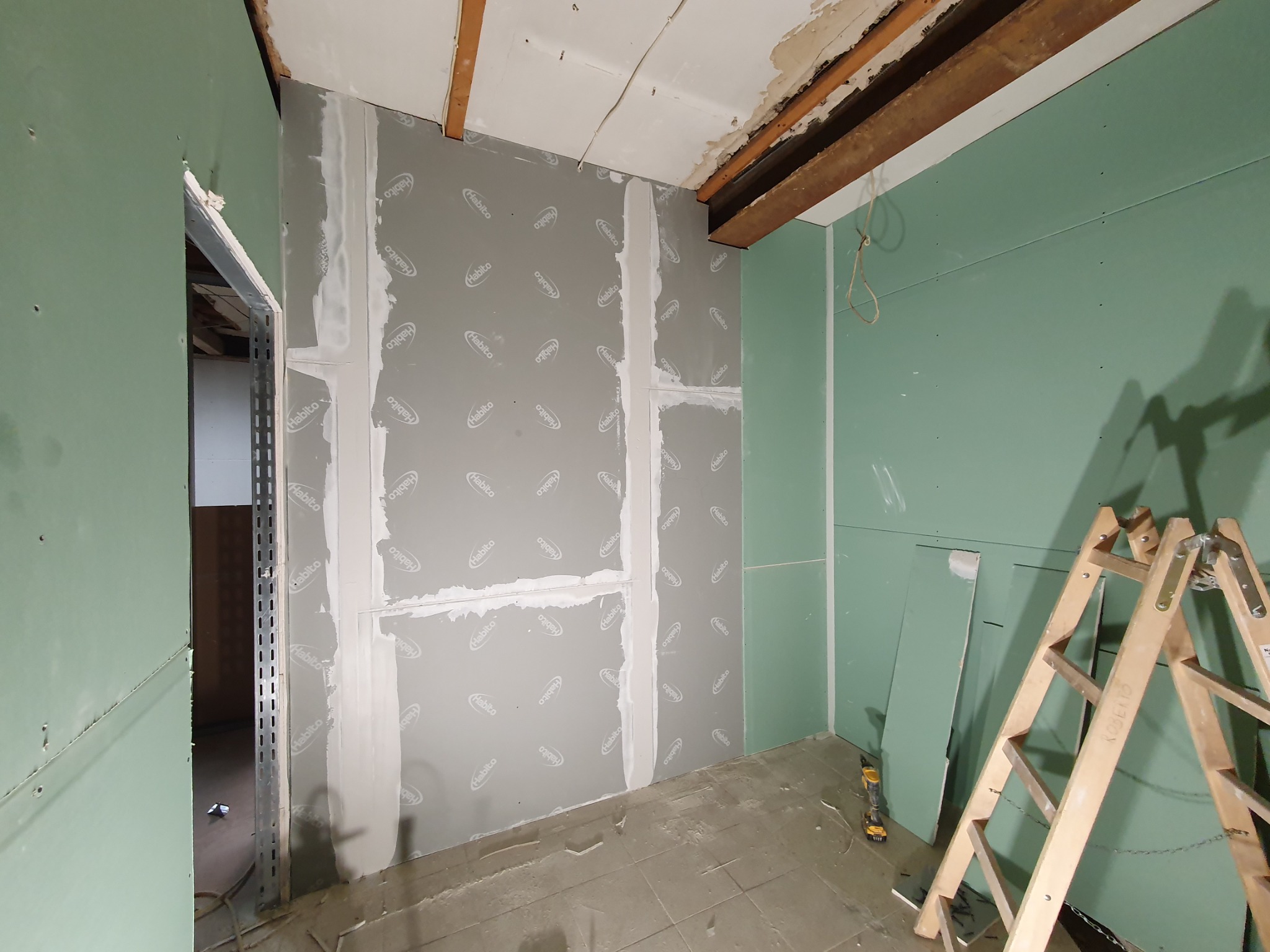 Wnętrze w trakcie prac wykończeniowych: szare i zielone ściany pokryte płytami z widocznym łączeniem, drewniana drabina oparta o ścianę, widoczne elementy konstrukcyjne stropu.