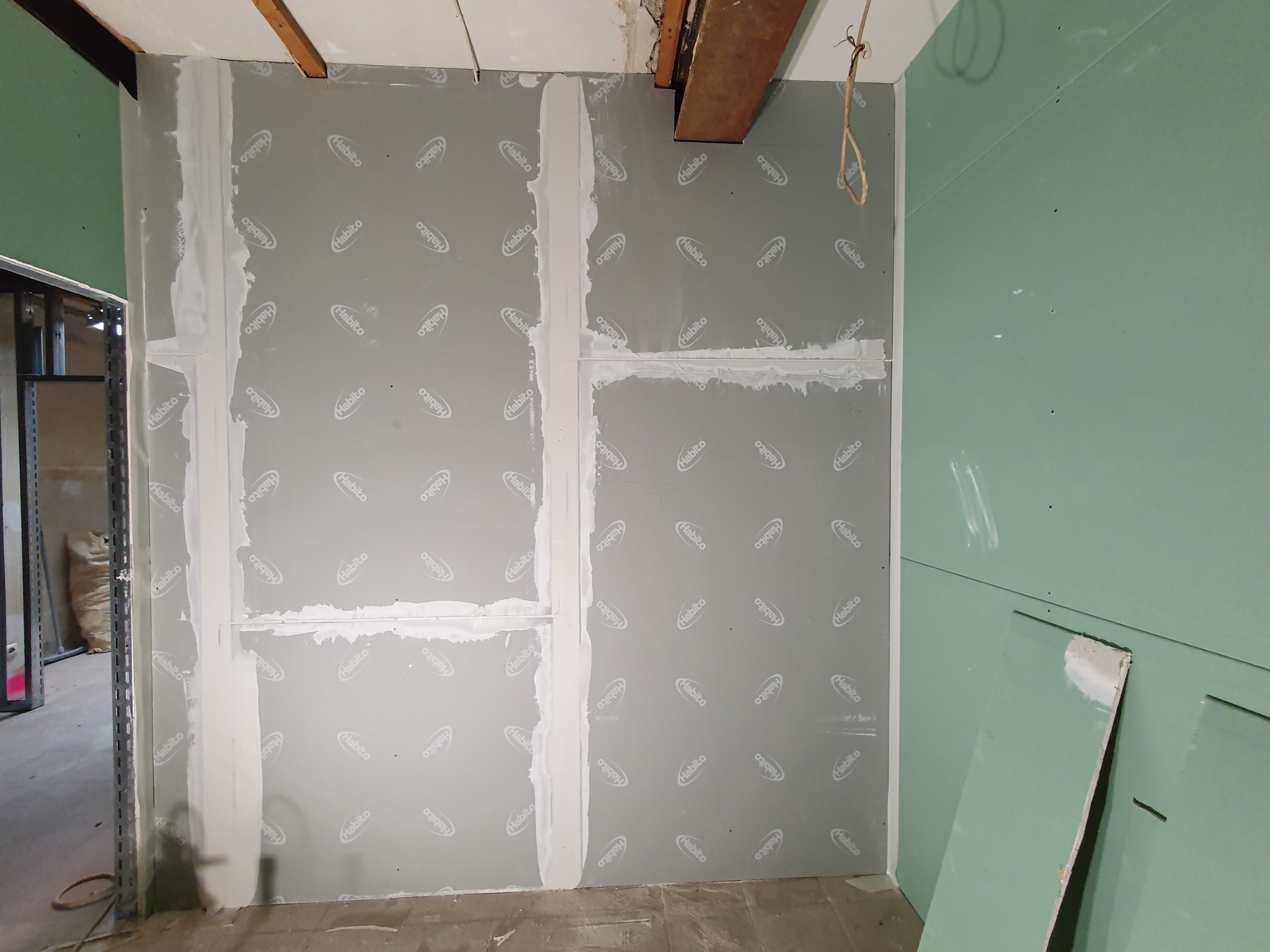 Ściana w trakcie montażu, pokryta szarą płytą z widocznym nadrukiem 'Habito', spoiny wypełnione gipsem, fragment sufitu z drewnianą belką, zielona ściana z boku.