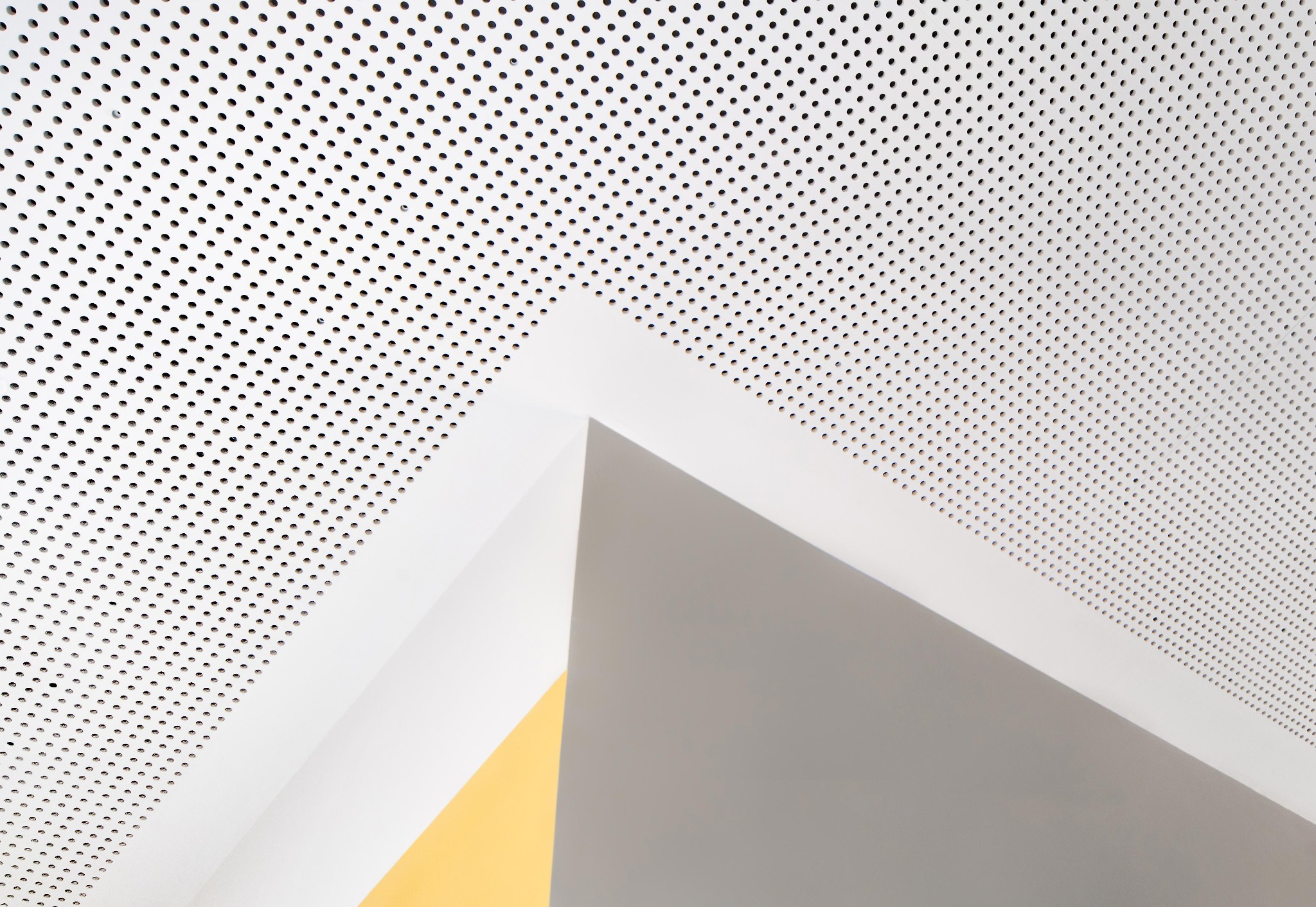 Perforowany sufit podwieszany z płyt w kolorze białym, widoczne fragmenty ścian w kolorze szarym i żółtym.