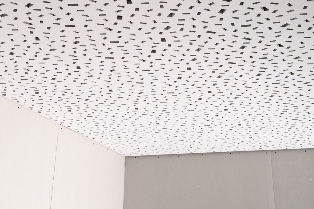 Biały sufit podwieszany z perforacją w kształcie nieregularnych prostokątów, zamontowany w narożniku pomieszczenia z widocznymi płytami gipsowo-kartonowymi na ścianach.