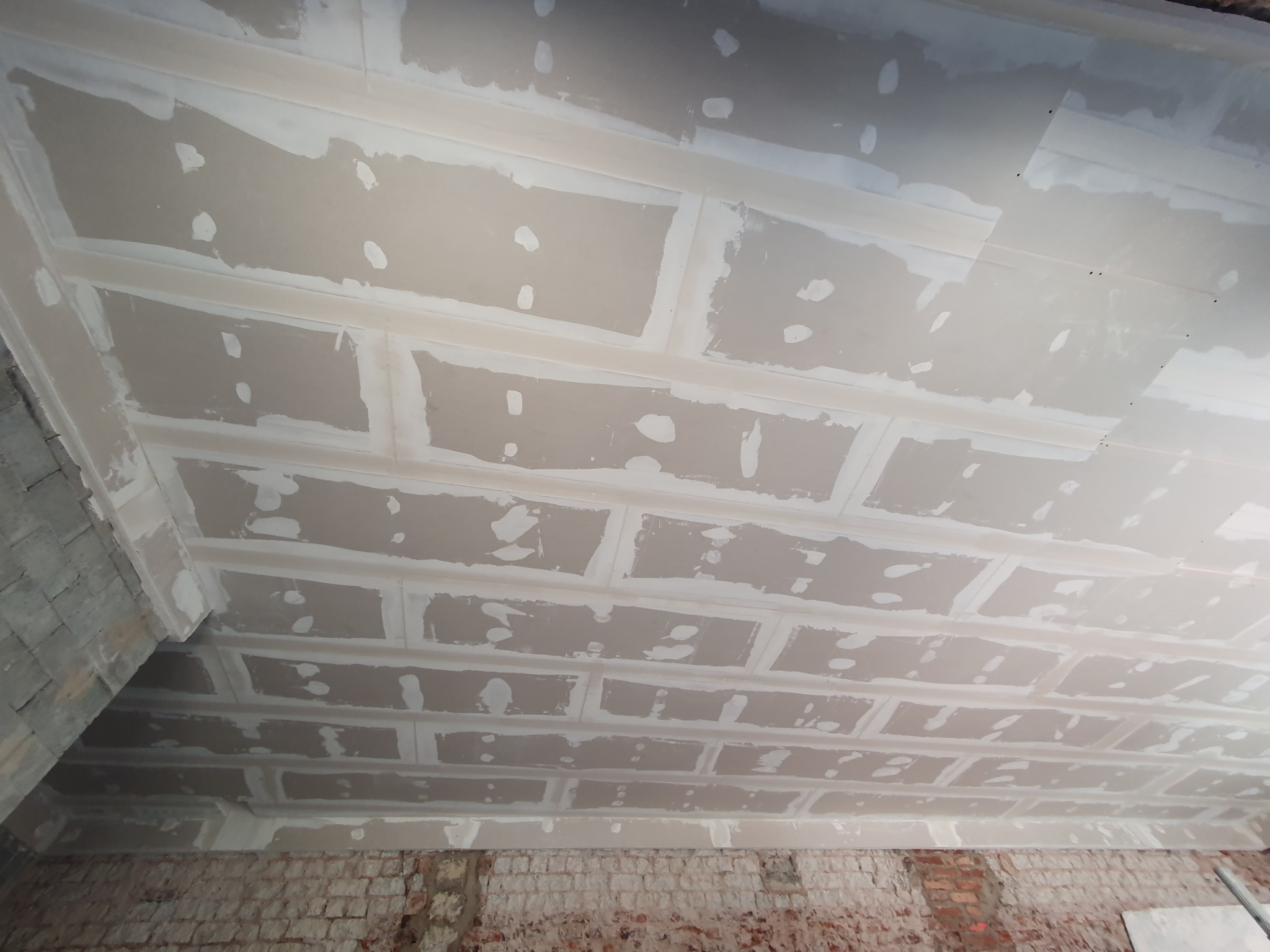 Sufit z płyt gipsowo-kartonowych w trakcie montażu, widoczne łączenia wypełnione masą szpachlową, surowa cegła w dolnej części ściany.