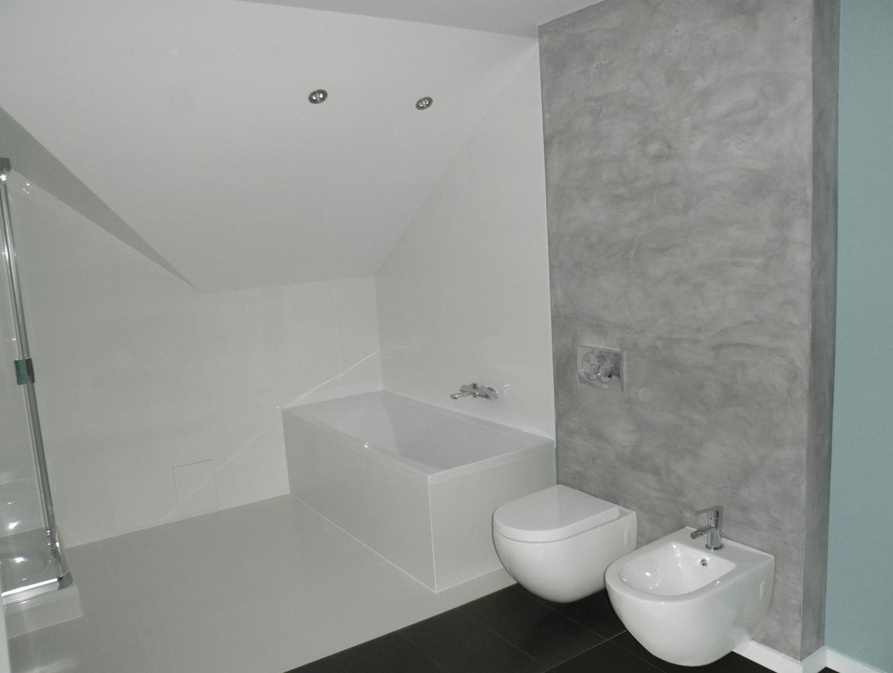 Nowoczesna łazienka na poddaszu z białą armaturą (wanna, toaleta, bidet) i szarą ścianą z efektem betonu architektonicznego, oświetlenie punktowe w suficie.