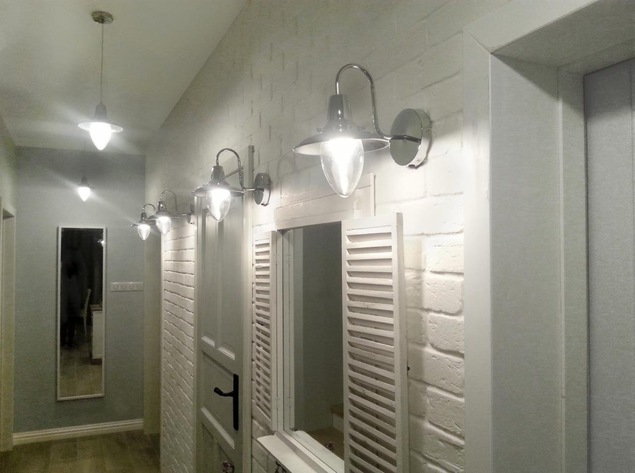 Korytarz w stylu rustykalnym z białymi ceglanymi ścianami, rzędem metalowych lamp ściennych z żarówkami w kształcie łezki i lustrem w białej ramie z okiennicami.