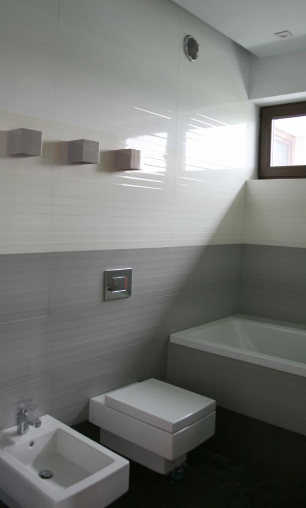 Nowoczesna łazienka z biało-szarą glazurą, podwieszaną toaletą, bidetem i wanną, oświetlona światłem wpadającym przez okno w drewnianej ramie.