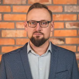 Portret mężczyzny w okularach i marynarce na tle ceglanej ściany, z widocznym znakiem wodnym fotografa w prawym górnym rogu.